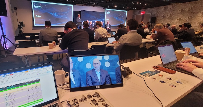 Attendees at a conference viewing a speaker presenting on a stage, while another participant watches a live feed of the event on their laptop screen in the foreground