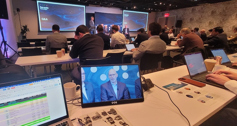 Attendees at a conference viewing a speaker presenting on a stage, while another participant watches a live feed of the event on their laptop screen in the foreground