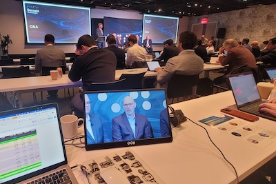 Attendees at a conference viewing a speaker presenting on a stage, while another participant watches a live feed of the event on their laptop screen in the foreground.