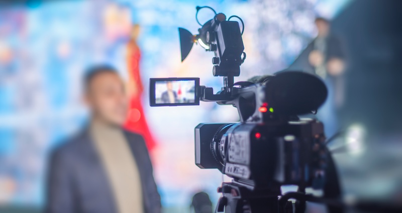Close-up of a professional video camera recording a person speaking on stage. The speaker and background are blurred, while the camera and its flip-out screen showing the subject are in sharp focus.