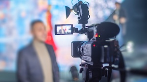 Close-up of a professional video camera recording a person speaking on stage. The speaker and background are blurred, while the camera and its flip-out screen showing the subject are in sharp focus.