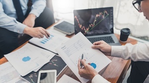 Two professionals review financial documents and charts at a table with a laptop displaying a rising stock graph, analyzing data and performance trends.