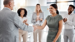 A senior executive congratulates a woman during a leadership announcement as colleagues applaud in the background during a workplace meeting.