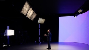 A presenter stands in a dark studio beside a large curved video wall, with production lights and a teleprompter screen visible during a video shoot.